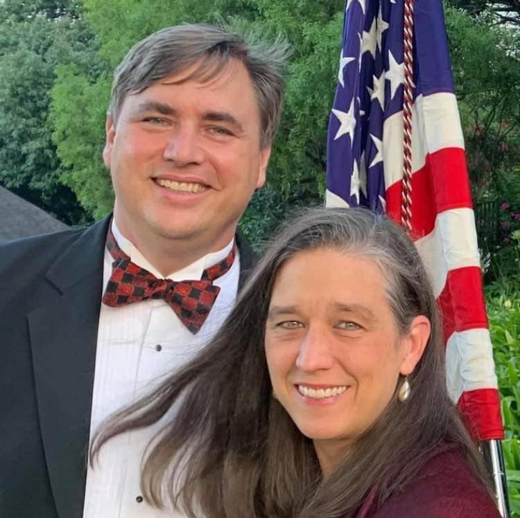 A couple poses together smiling, with an American flag in the background.