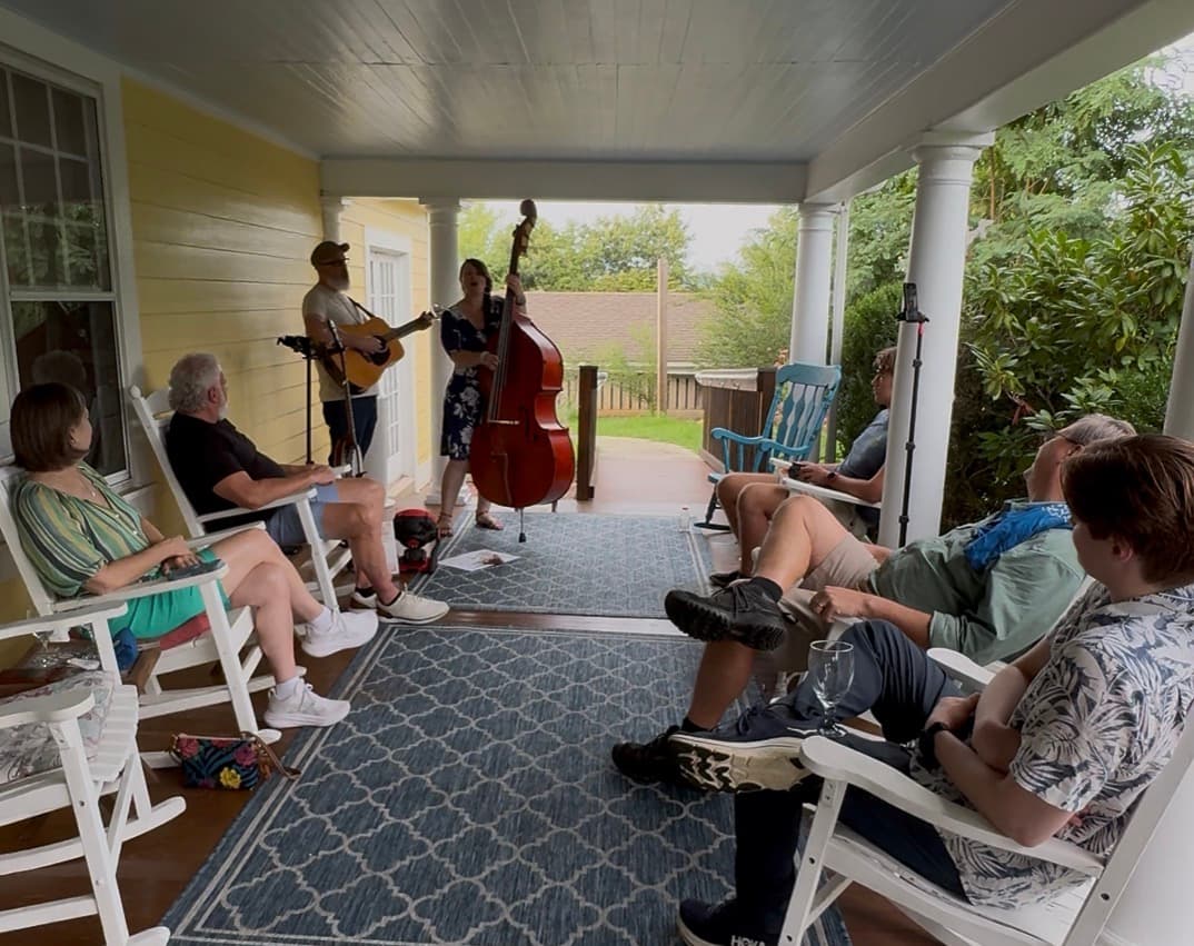 A musician plays guitar while a singer performs with a double bass, entertaining a small audience on a porch.