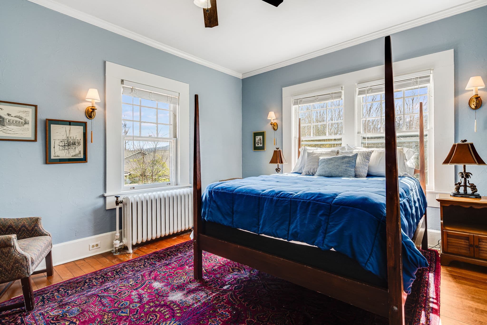 queen bed with blue bedspread, bedside lamps, windows, chair