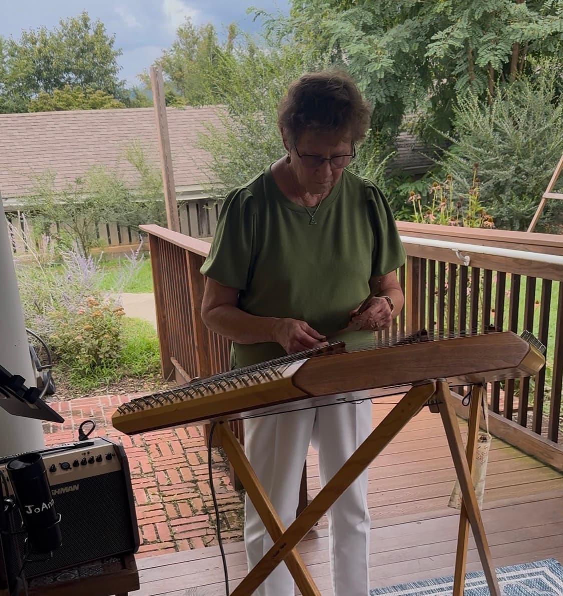 An older woman plays a zither on a wooden deck surrounded by greenery.