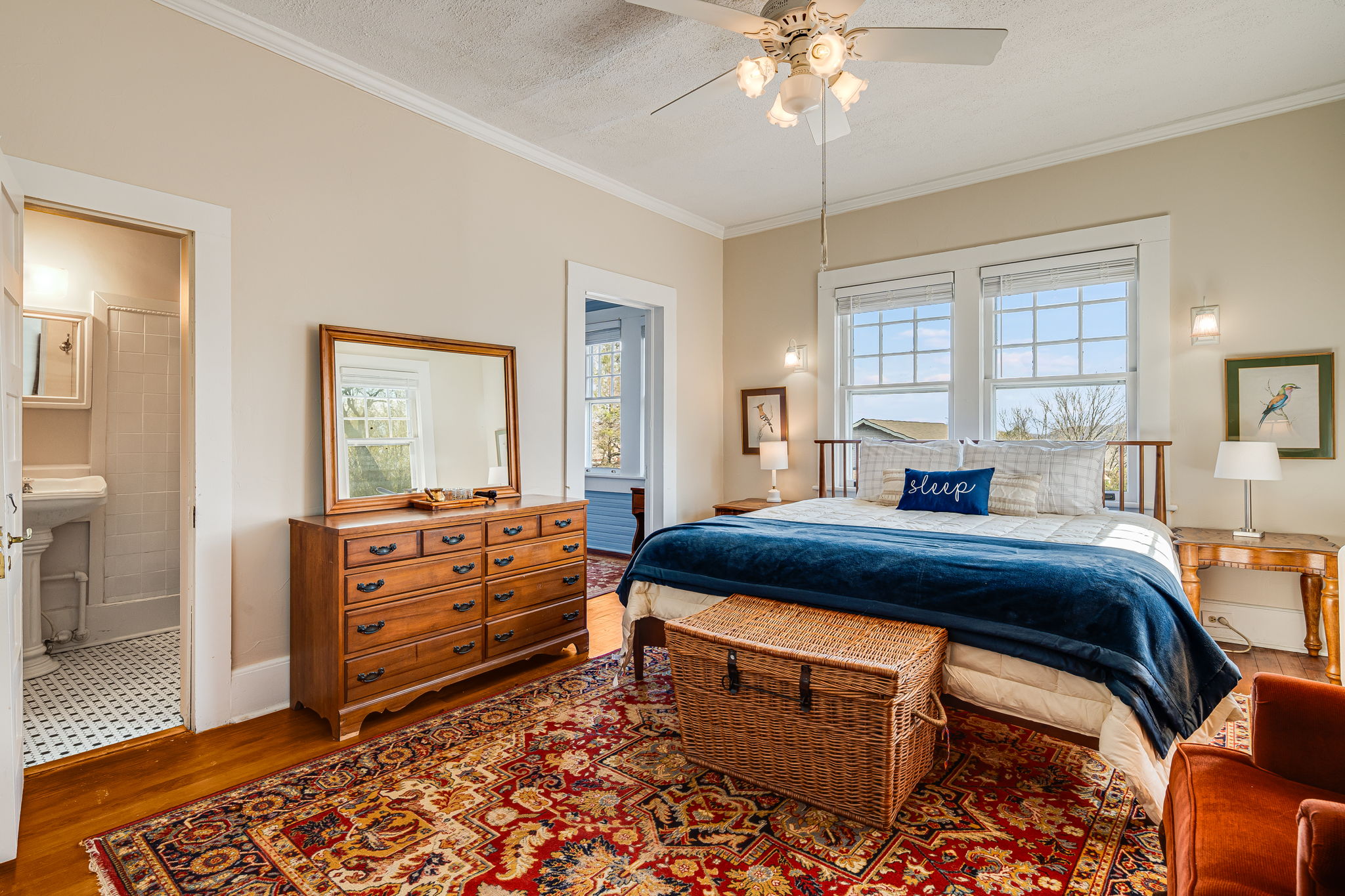 king bed, oriental carpet, blue bedspread, two windows, bedside lamps, dresser, bathroom