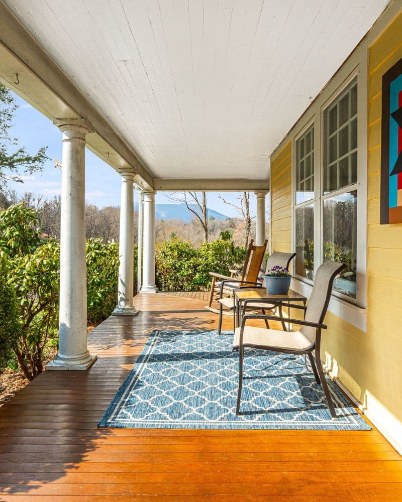A cozy porch featuring two chairs and a decorative rug, framed by columns and surrounded by greenery.