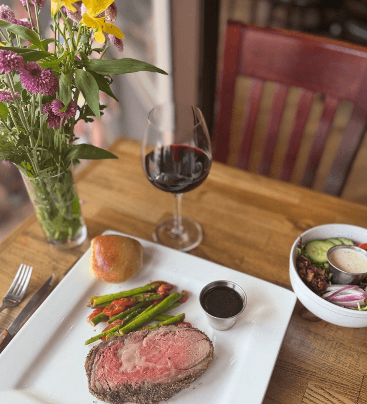 A plate with sliced roast beef, asparagus, a roll, dipping sauce, a salad, and a glass of red wine sits on a wooden table alongside a vase of flowers.