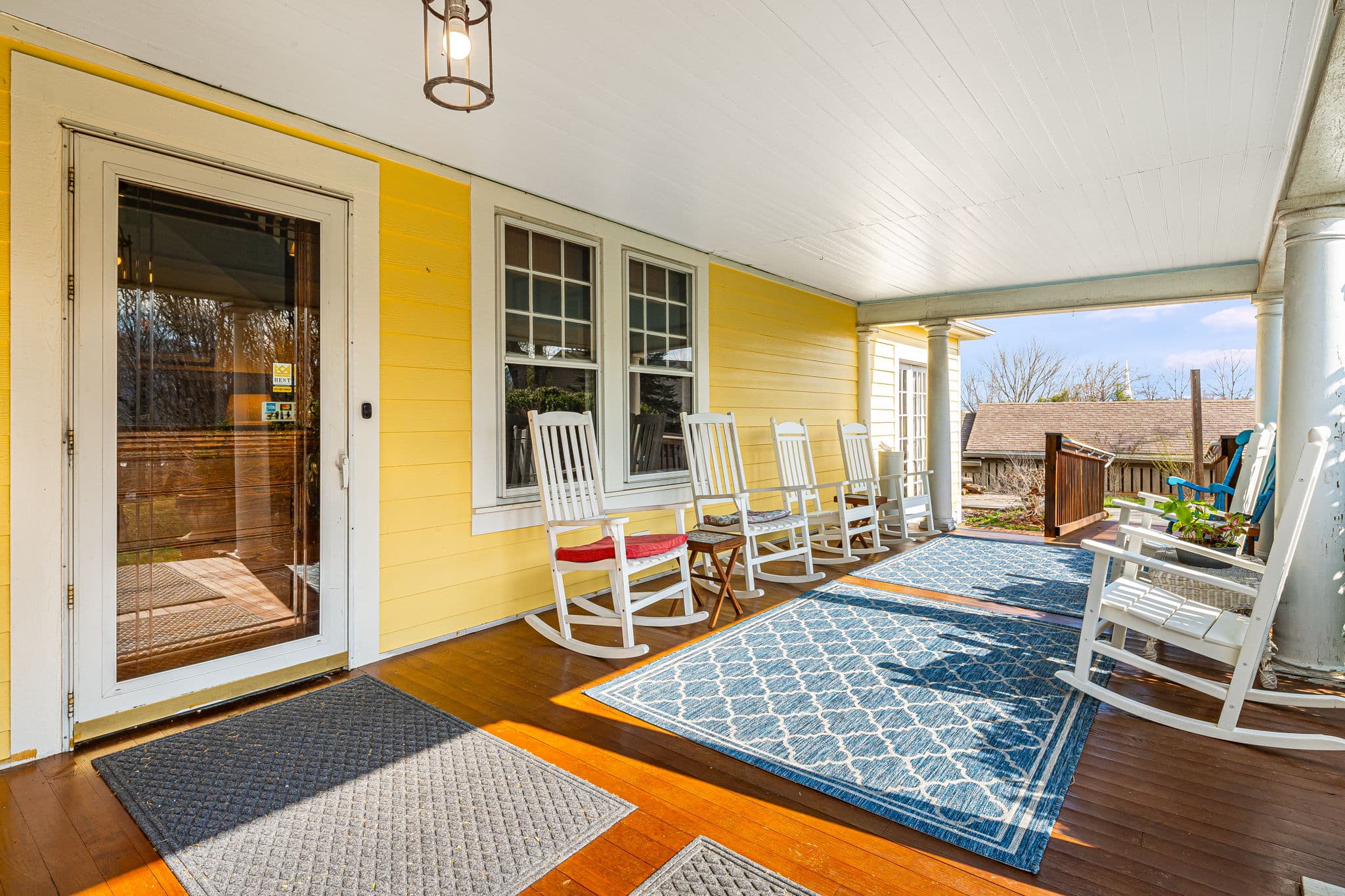 large porch with white rocking chairs on blue rug, windows, door, overhead light, blue skies