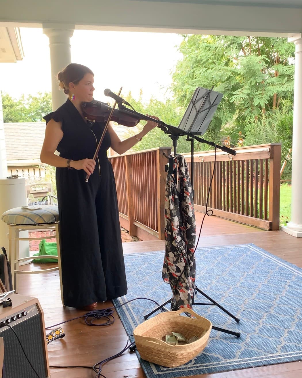 A woman in a black jumpsuit plays the violin on a porch, with music sheets and a decorative basket nearby.