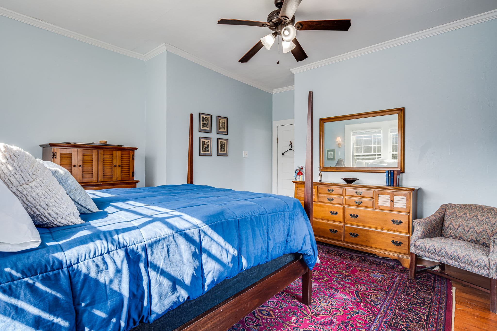 queen bed with blue bedspread, large mirror, ceiling fan, chair, two dresers