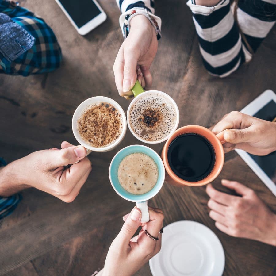 Four hands holding different coffee cups gathered together on a wooden table.