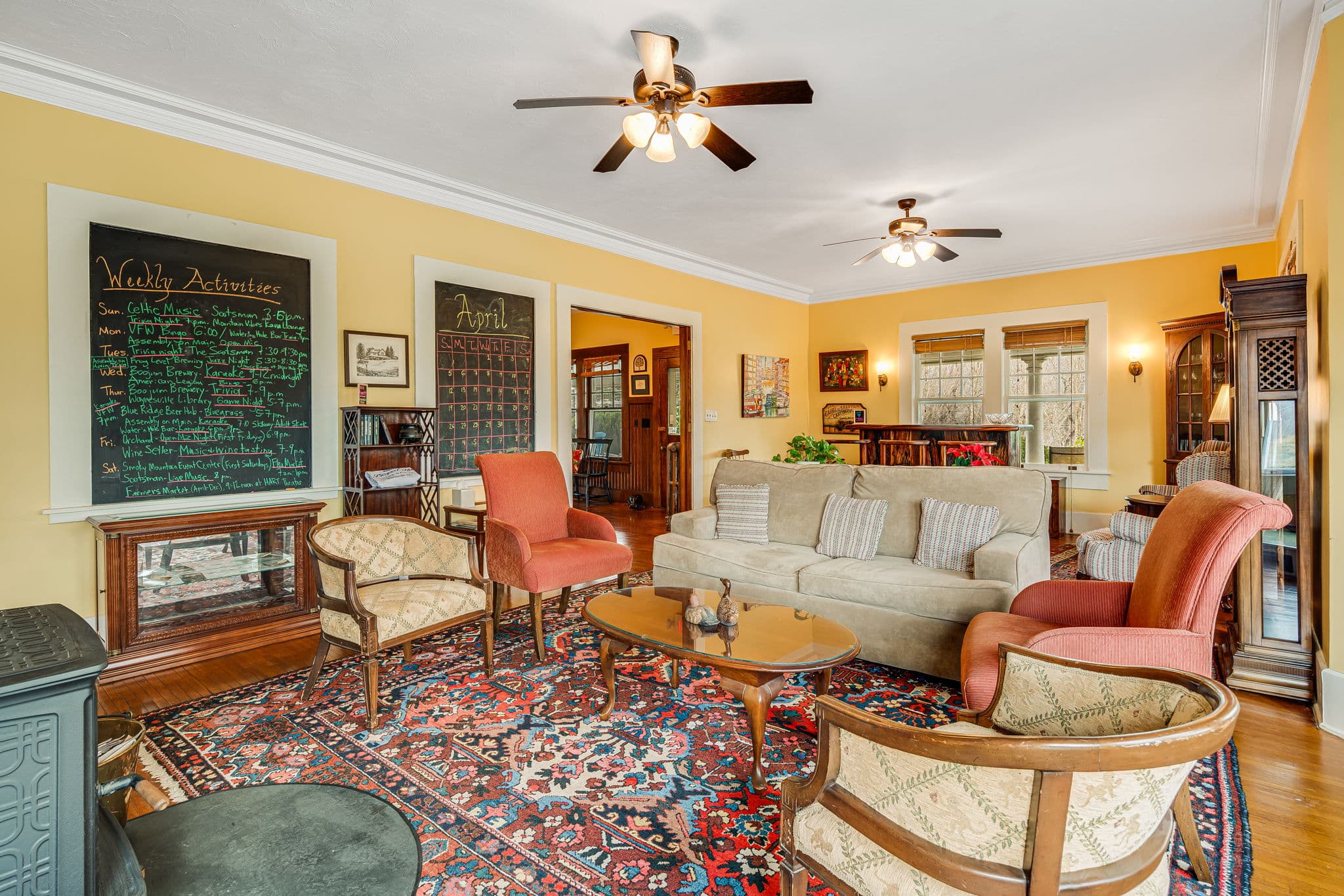 parlor or living room, fireplace with wood stove, two large windows behind South African wooden bar, grandfather clock, four chairs, large sofa, coffee table, oriental carpets, doorway to foyer or entrance, chalk boards listing local activities