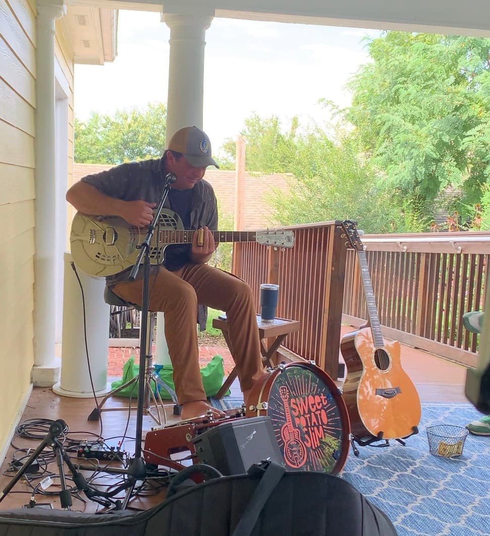 A musician plays slide guitar on a porch, surrounded by instruments and greenery.