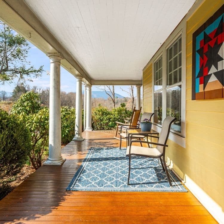 A sunny porch featuring wooden flooring, two chairs, a decorative rug, and vibrant greenery.
