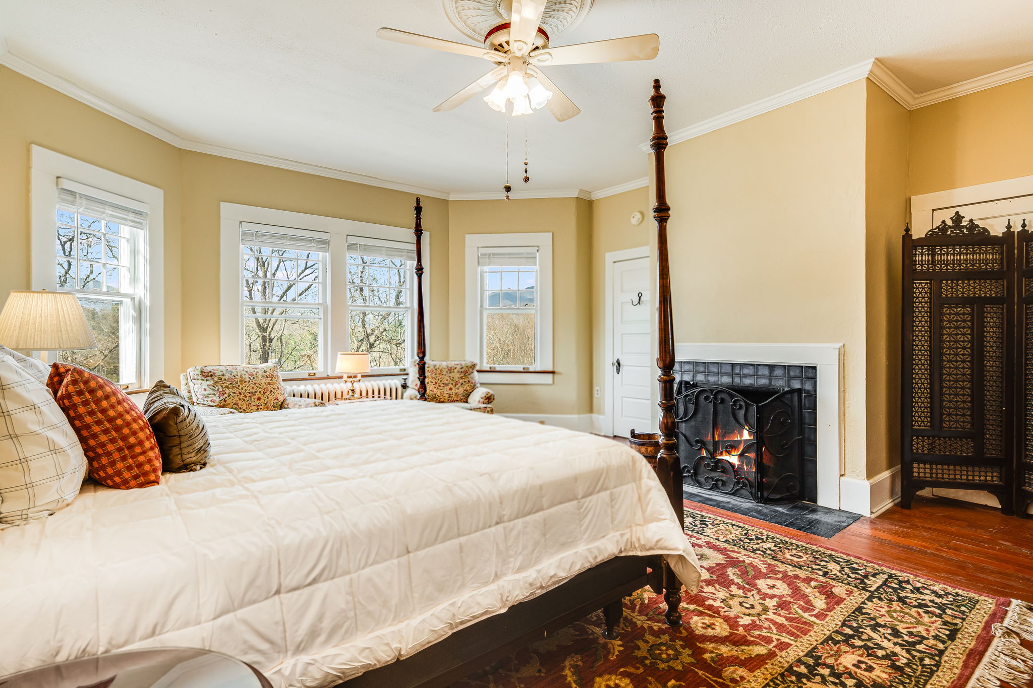 king bed, ceiling fan, oriental carpet, white bedspread, pillows, three windows.