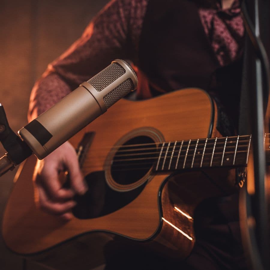 A close-up of a musician's hand playing an acoustic guitar with a microphone in the foreground.