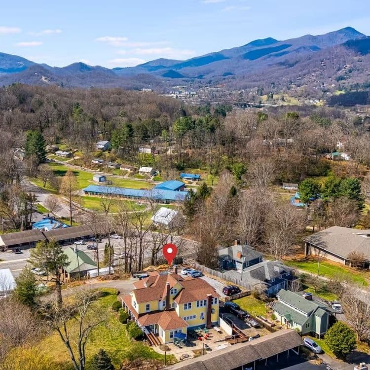 Aerial view of a colorful house surrounded by trees and mountains, with small buildings and roads in the vicinity.