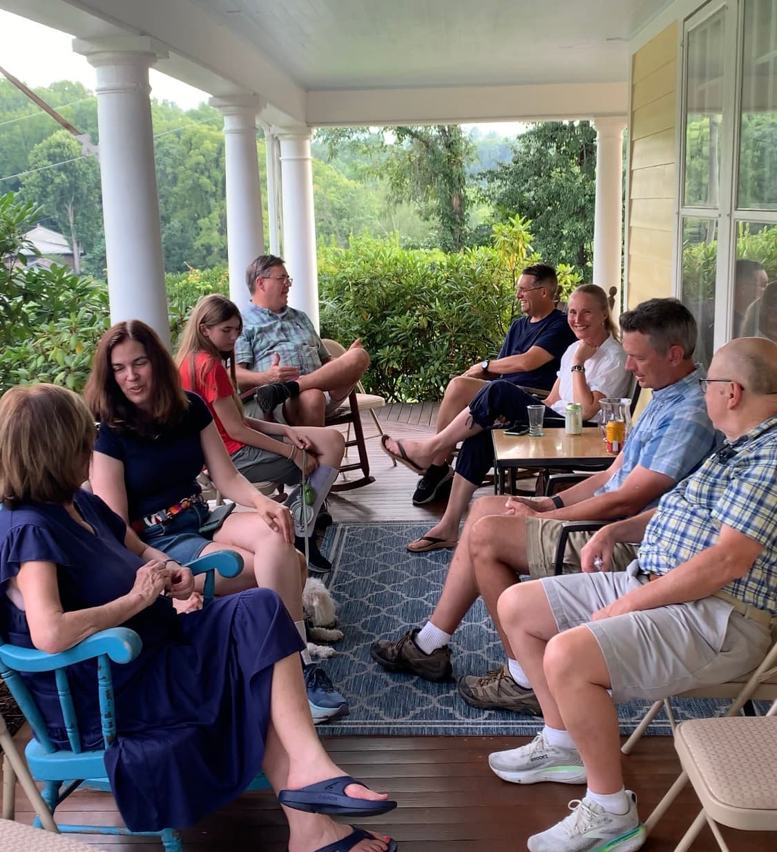 A group of people gathers on a porch, enjoying conversation in a lush, green setting.