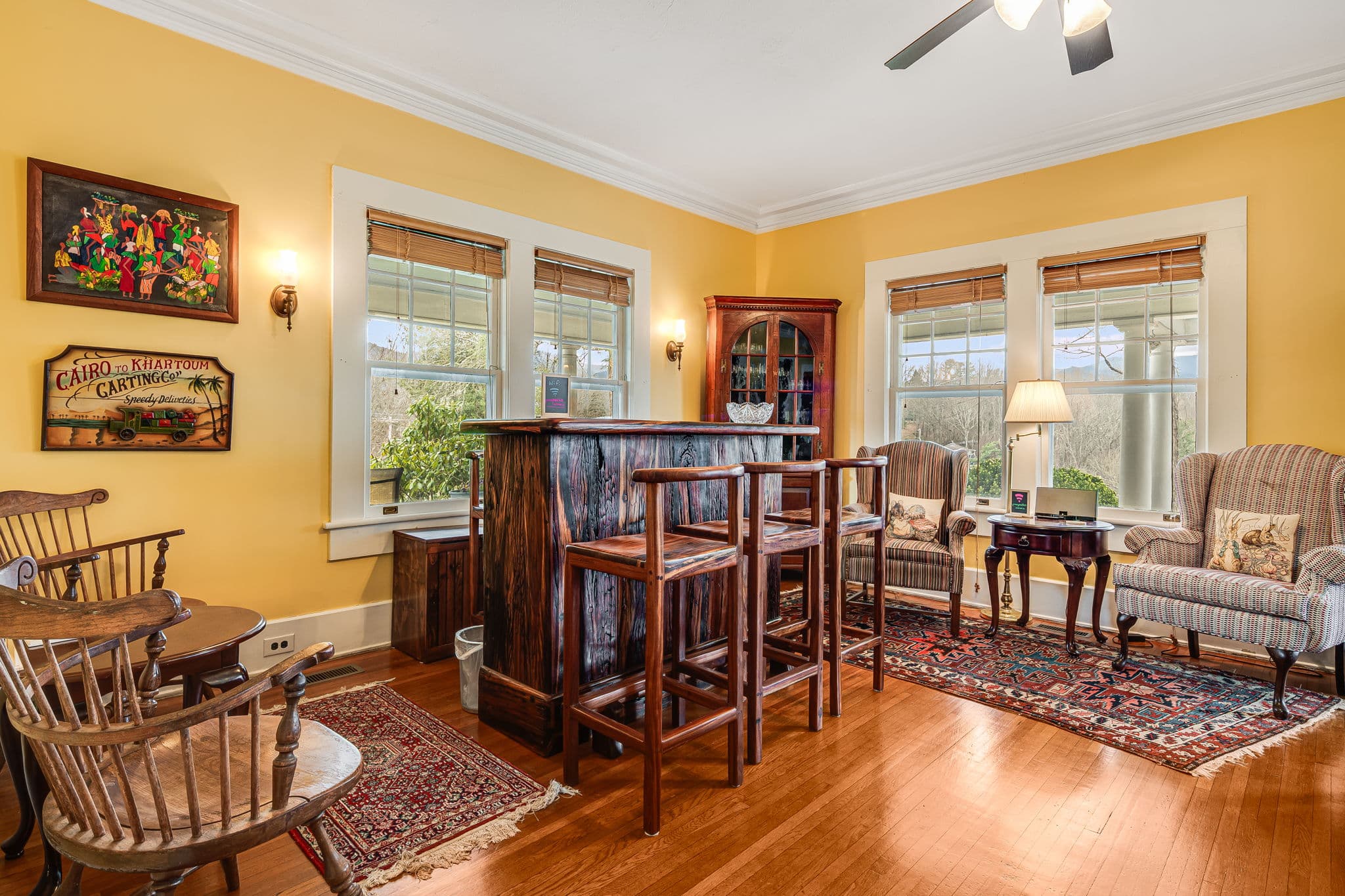 Wooden South African bar with barstools, four chairs, side tables, oriental carpets, African paintings, antique corner cabinet, four windows, ceiling fan