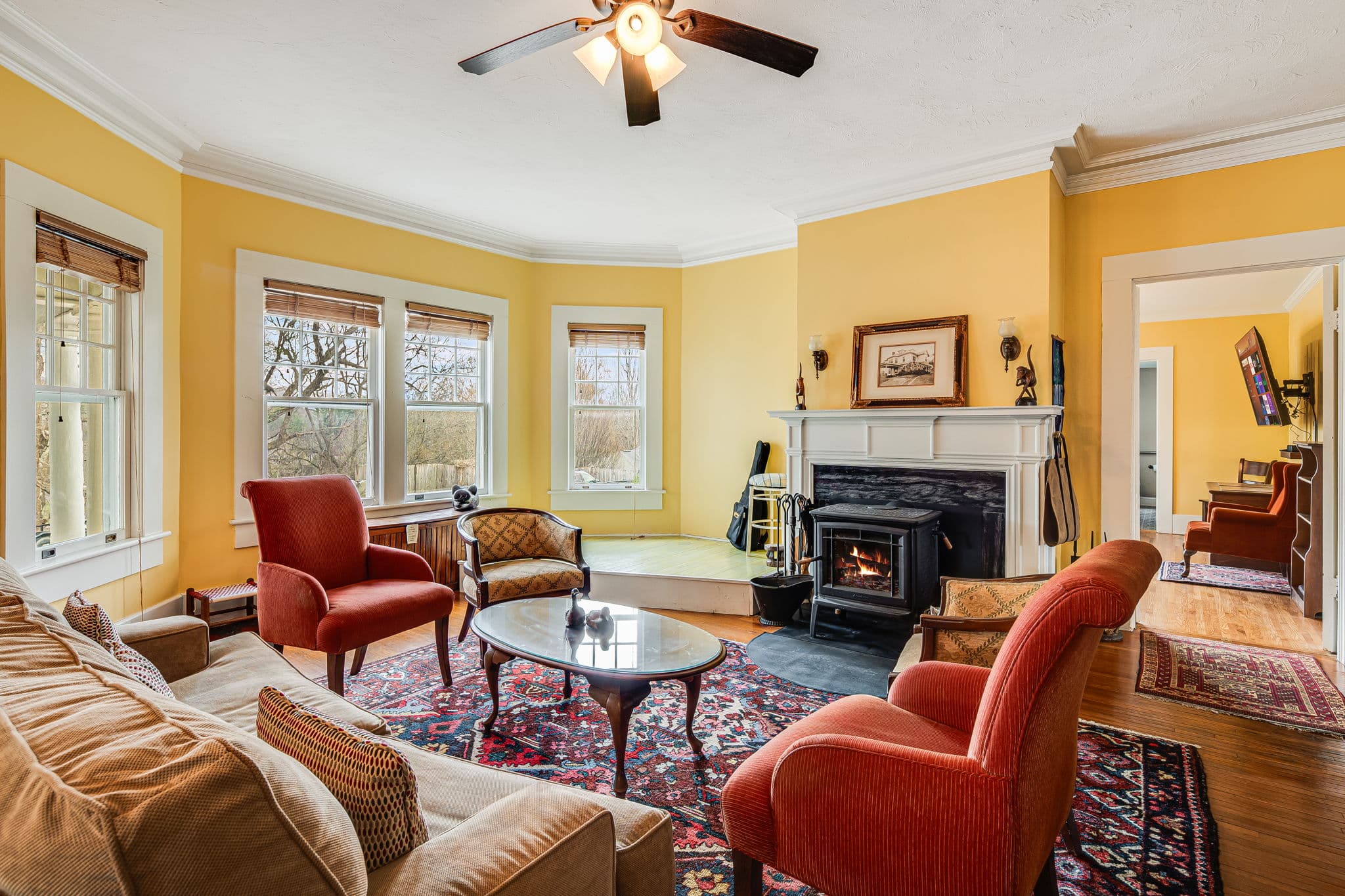 parlor or living room, fireplace with wood stove, antique photo of historic inn, four large windows, four chairs, large sofa, coffee table, oriental carpets, mantle with wooden carved monkeys, doorway to tv room.