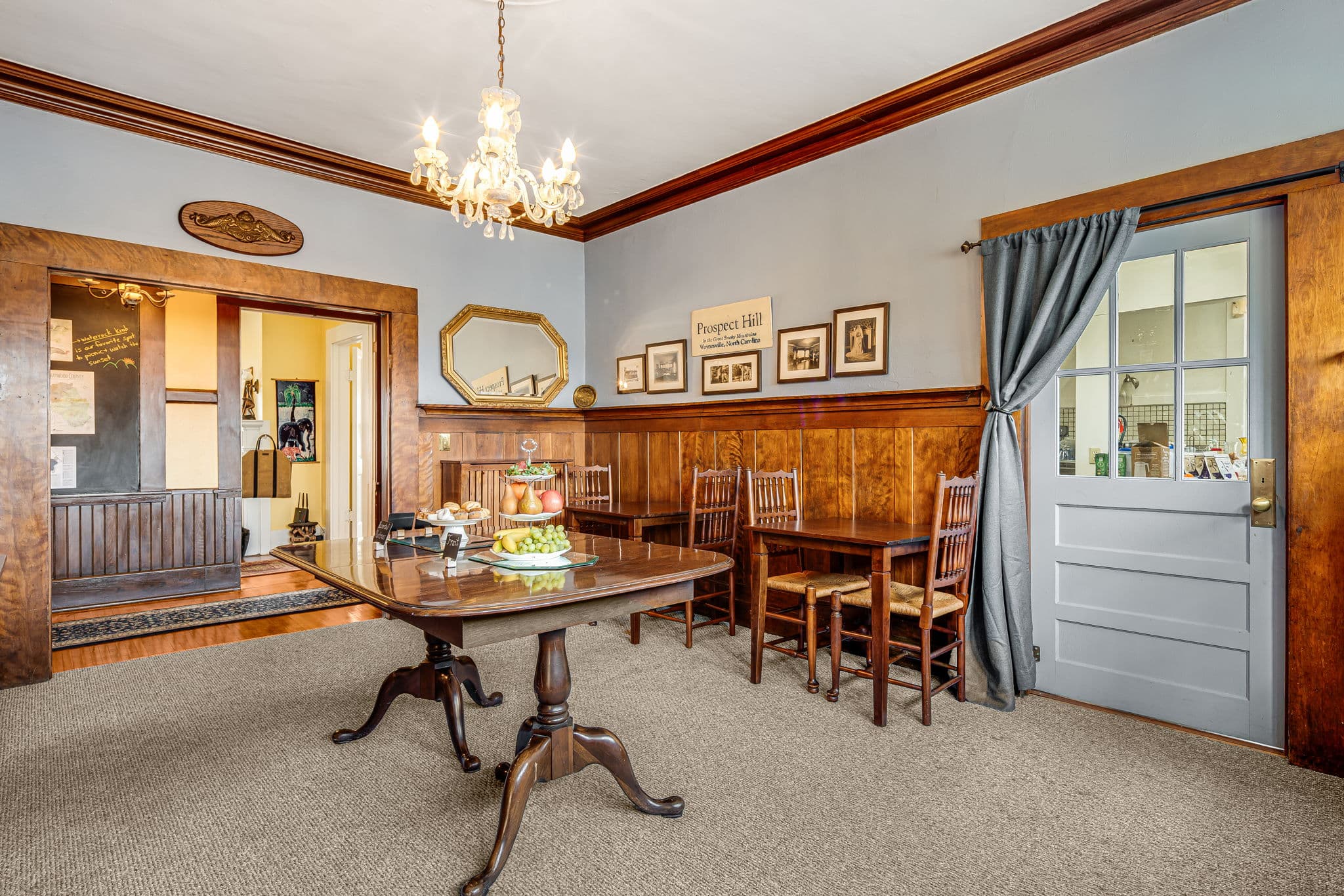 dining room with tables and chairs, historic photos and memorabilia on wall, mirror, entrance to commercial kitchen, entrance to hallway to foyer, crystal chandelier